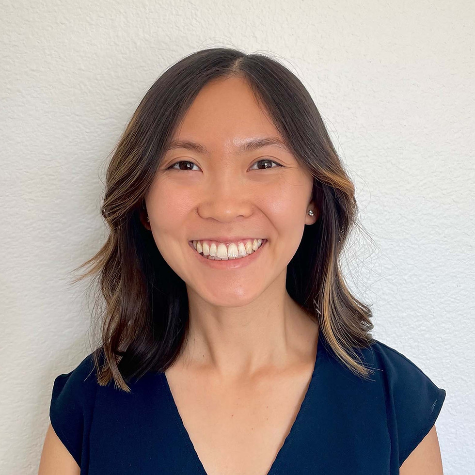 An Asian-American woman in her 20s-30s with fair to medium skin, dark brown eyes, and straight black to medium brown hair with light highlights. She smiles widely, wearing a dark blue V-neck blouse and a round earring, with a light-colored wall in the background.