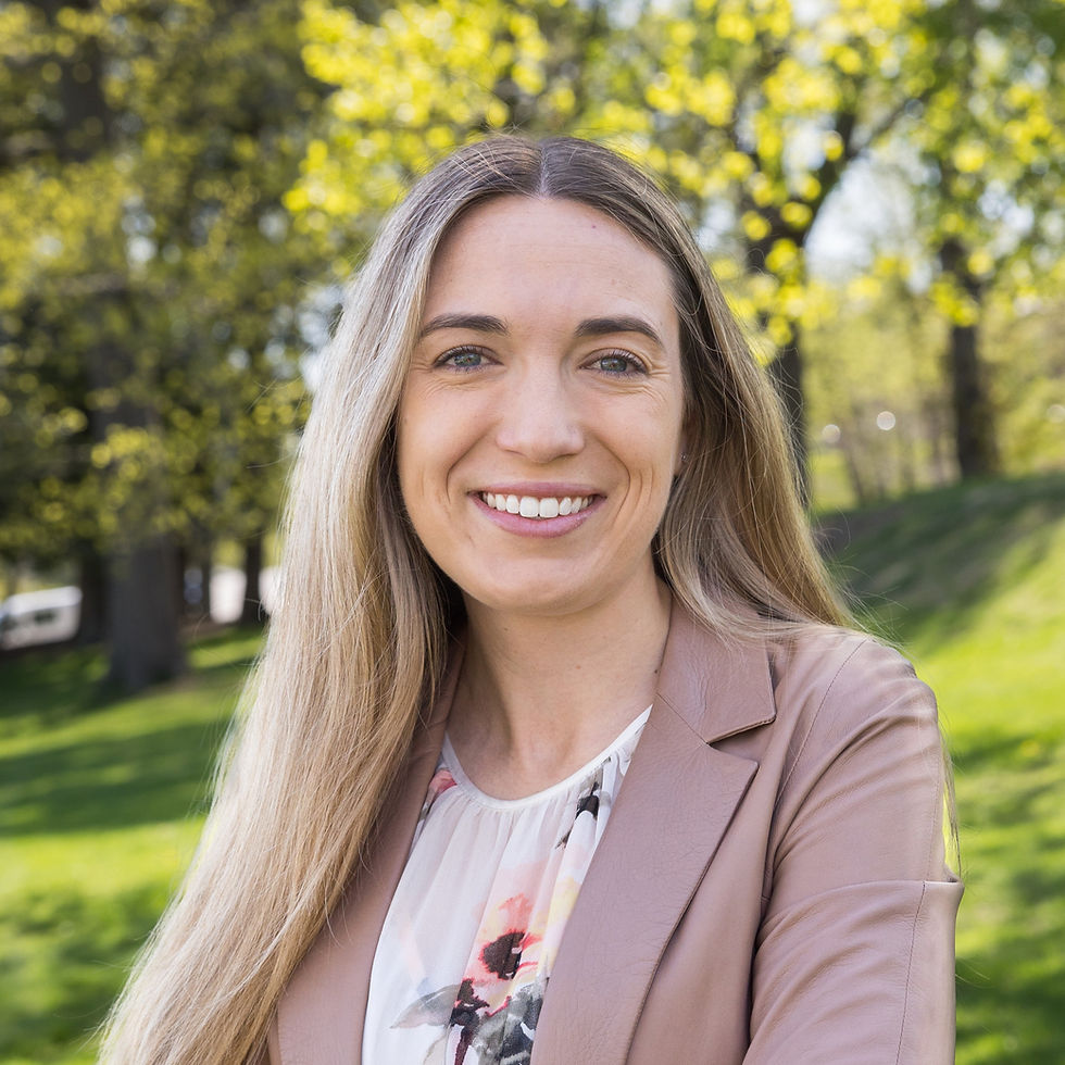 An American woman in her early 30s with medium skin, blonde straight hair, and blue eyes, smiling. She wears a brown jacket and white floral blouse, standing in the sunlight with a blurred natural background.