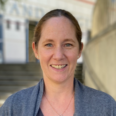 A European woman aged 40–50 with fair skin and brown hair parted in the middle and tied back. She has green eyes, well-shaped eyebrows, and a gentle smile. She wears a gray, long-sleeved open-front jacket over a black shirt, along with small gold earrings and a matching necklace. She faces forward, with a brownish-gray staircase and a white building with blue doors in the background.