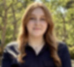 An American woman in her 20s with fair skin and long, curly blonde hair that creates the impression of a flowing look. She has blue eyes highlighted by subtle makeup, matching well-groomed eyebrows, and a soft smile. She wears a black V-necked sweater and stands in front of a green, foresty background.