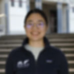 An Asian/South American woman in her 30s with fair skin and dark brown hair parted in the middle and tied in a low ponytail. She has dark brown eyes and wears thin, round black-framed glasses. She is wearing a long-sleeved blue jacket with a front zipper, featuring the Tarling-Vallim lab logos, over a light-colored shirt. She faces forward, and in the background, there is a brownish-gray staircase with a railing leading to a property with white walls and doors and details.