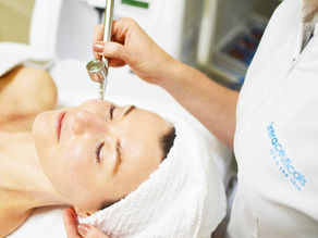 Woman receiving a facial treatment with an Intraceuticals pressurized oxygen device, wrapped in a white towel. Medical esthetician wears a white coat with "intraceuticals" text.