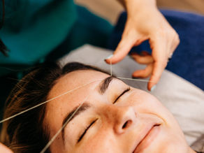 Close-up of a woman's face during eyebrow threading. Hands hold thread over the eyebrows. Her eyes are closed, implying relaxation.