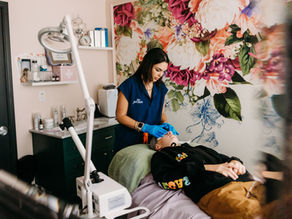 A woman in a blue shirt with "The Parlour" text gives a facial treatment, emitting steam. Background has a floral mural and shelves. The medical spa is The Parlour located in Pinecrest, Miami, Florida, near South Miami, Palmetto Bay, and East Kendall. Shelves with skincare items visible in the background. The Parlour specializes in facial treatments, chemical peels, anti-aging injection treatments by a licensed nurse practitioner, laser hair removals, brow shaping, and more.