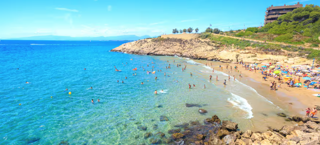Crowded sunny beach with swimmers in turquoise water, colorful umbrellas on the sand, rocky cliffs, and a calm seaside mood.