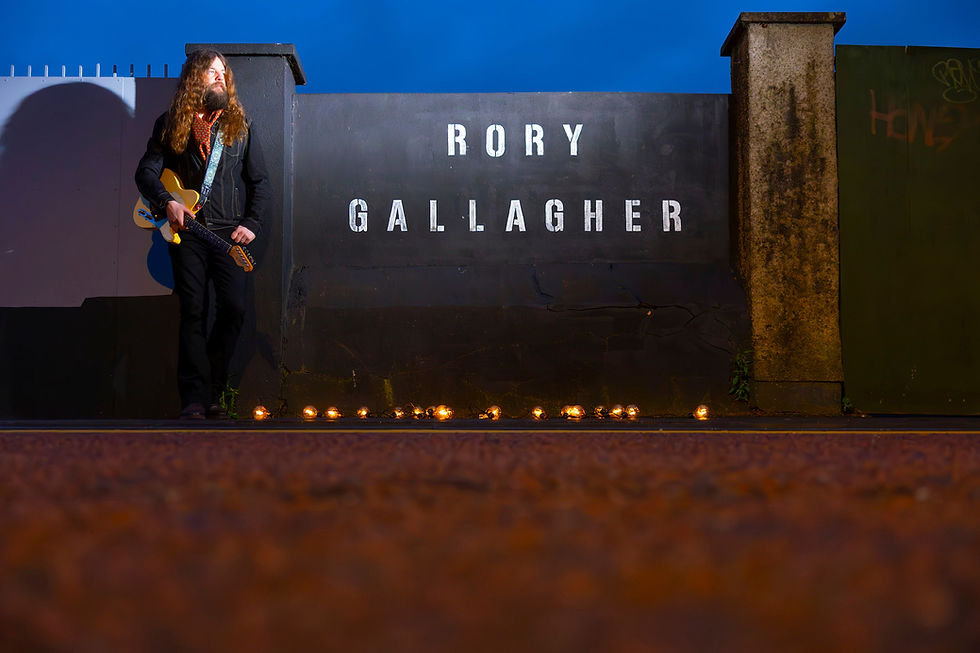 Man with guitar leans against wall with "Rory Gallagher" text. Candle lights on ground create a warm ambiance. Blue sky in background. Cork Jazz fest