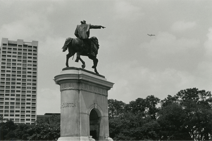 Black and white image of statue pointing at a plane. 