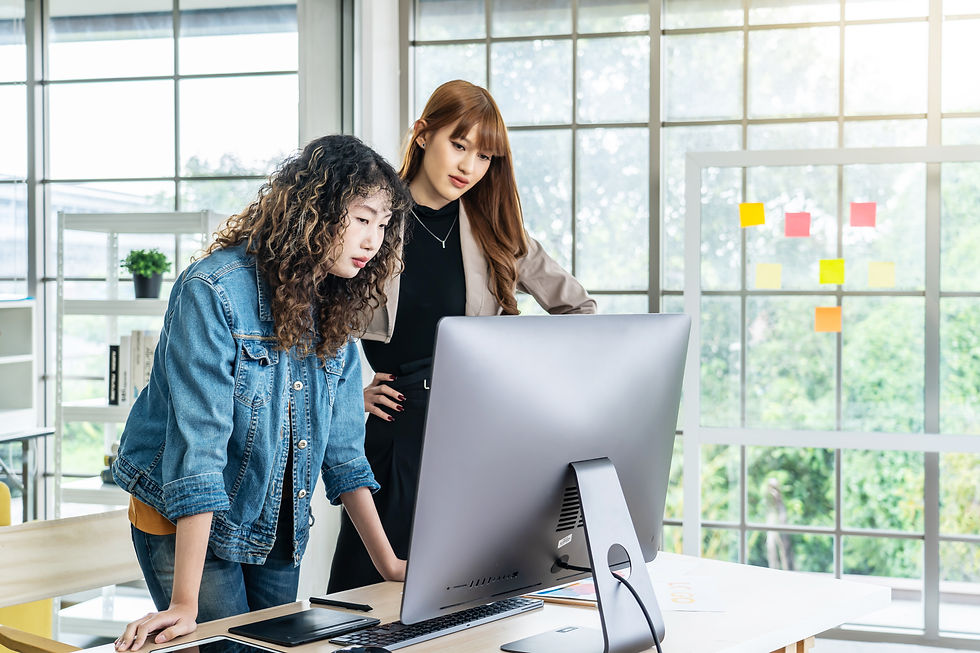 Two women focus on a computer screen in a bright office, with sticky notes on a window. One stands with hands on hips, the other leans forward.