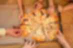 Hands reaching for pizza slices in an open box on a wooden table. The pizza has toppings like olives and sausages, creating a shared, joyful moment.