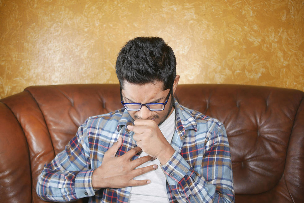 Man in plaid shirt and glasses sits on brown couch, hand on chest and coughing. Yellow textured wall in the background. Appears unwell.