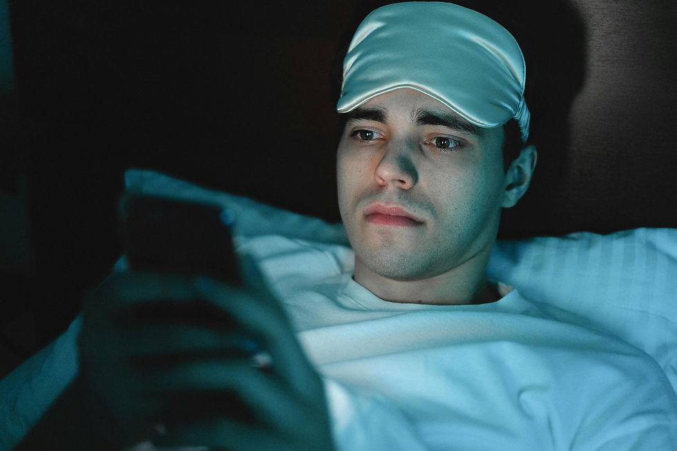 Man in bed wearing a sleep mask, looking at a phone with a concerned expression. Dim, blue-lit room, suggesting nighttime.