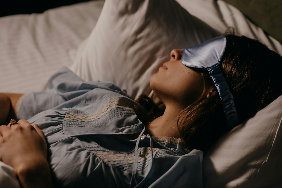 Woman in light blue pajamas, wearing a blue sleep mask, lying on white pillows, resting in a calm, dimly lit bedroom setting.
