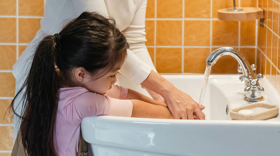 Woman helps girl wash hands at sink with running water. Background has orange tiles. Calm, instructional moment.
