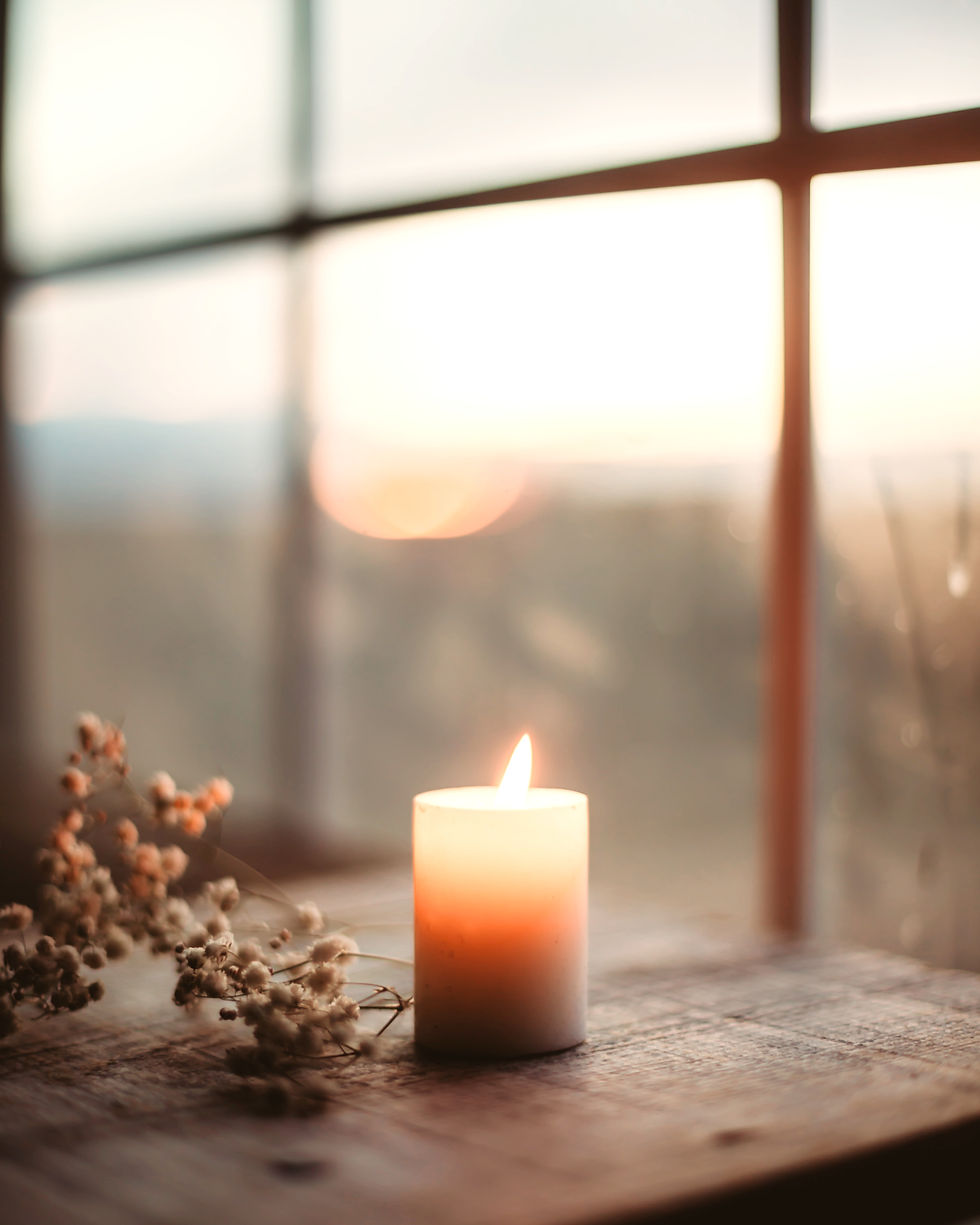 Une bougie allumée placée devant une fenêtre, accompagnée d’une petite fleur séchée, baignée d’une lumière douce du matin ou du soir, créant une ambiance calme et introspective.