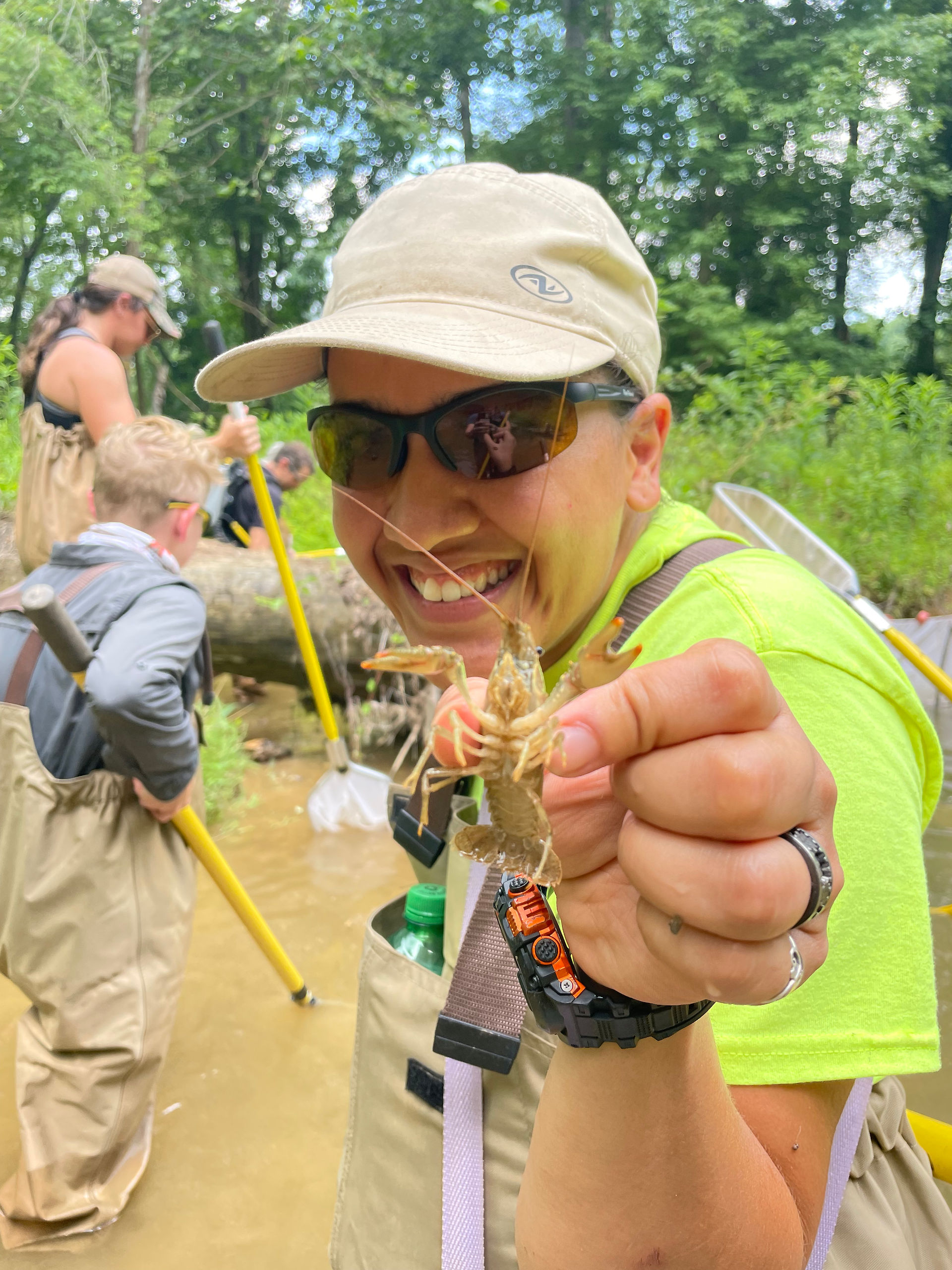 Crew, Xai, holding a crawfish outwards towards viewer while smiling, wearing sunglasses and a hat