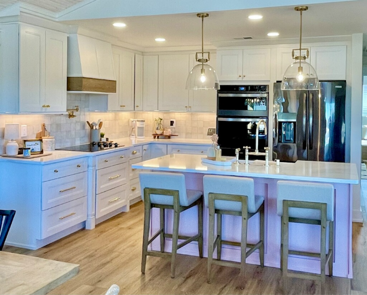 Bright, modern kitchen with white cabinets, island, and barstools. Pendant lights hang above. Black double oven and fridge are visible.