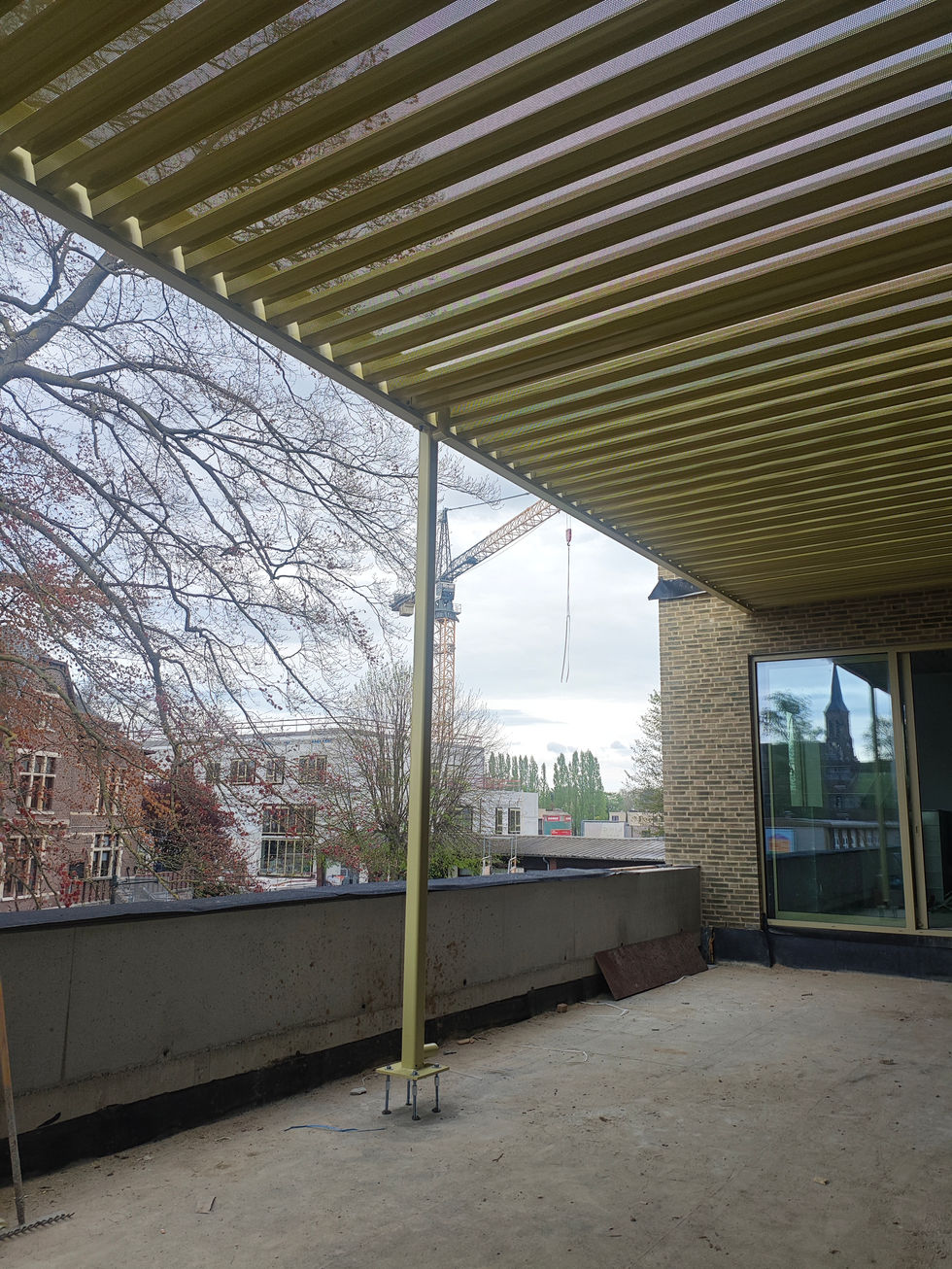 De eerste luifel over het terras op niveau +1 is geplaatst—een geperforeerde staalplaat in galva met een zachte champagnekleur die het zonlicht filtert tot een spel van stippen en schaduwen. De lichte constructie zweeft boven het terras als een elegante kap, functioneel en verfijnd tegelijk. Onder de perforaties vang je in de verte een glimp van het kasteel en het in opbouw zijnde atheneum: een subtiel perspectief dat heden, verleden en toekomst in één beeld verbindt. Deze luifel is meer dan beschutting—het is een gebaar van architecturale gastvrijheid, licht en luchtig, maar met een sterke aanwezigheid.