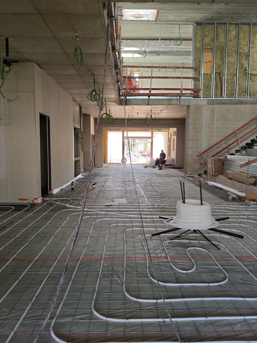Na de verdieping slinger een dans van witte leidingen nu ook over het gelijkvloers—de vloerverwarming wordt gelegd. Als een fijnmazig netwerk tekenen ze zich af tegen de kale betonvloer, klaar om straks onzichtbaar warmte te verspreiden in de klaslokalen en gangen.