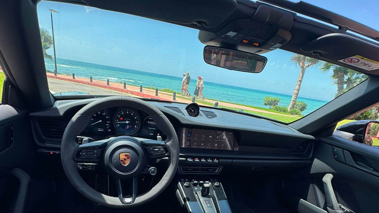 Interior dashboard and steering wheel of a Porsche 911 Targa 4 GTS with panoramic sea views through the open roof.