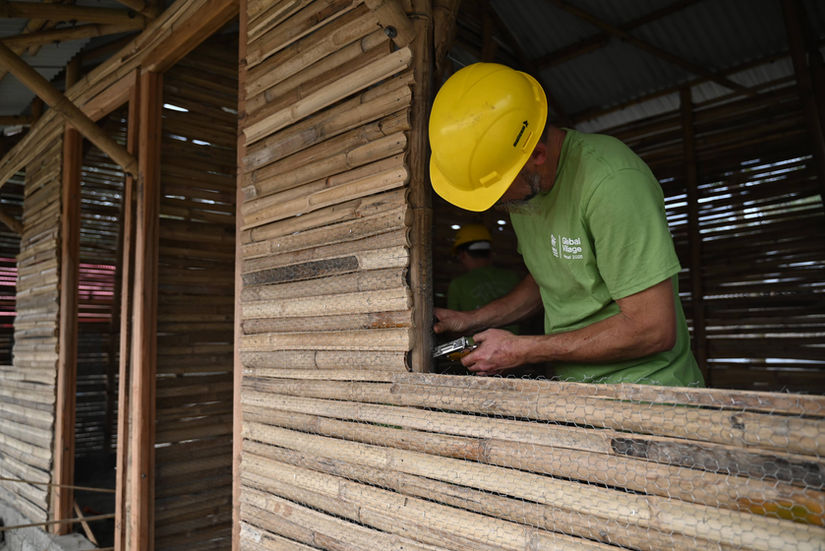 A man in a yellow hard hat in front of a bamboo home under construction. He holds a staple gun.