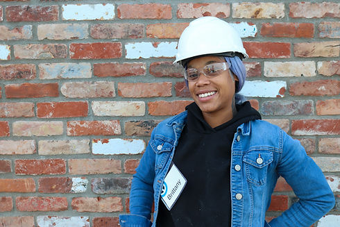 Young woman in a hard hat and safety glasses standing in front of a brick wall