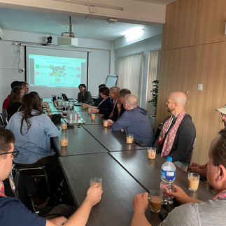 Group of people sitting around a conference table. A woman is presenting a presentation about Habitat Nepal at the front of the room.