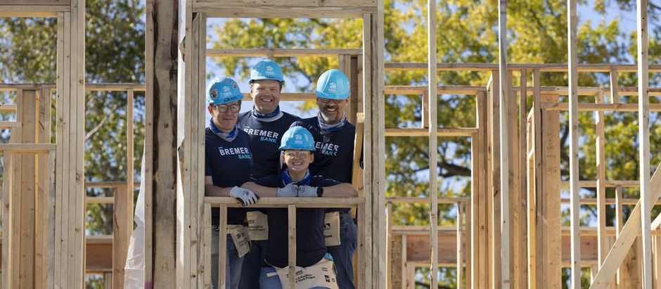 A group of people looking through the space where a window will go on a framed house under construction. The group are all wearing blue Habitat for Humanity hard hats and matching navy shirts.