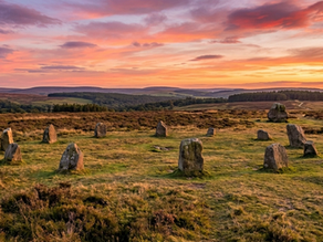 Nine Ladies stone circle on grass under a vibrant sunset sky; rolling hills and forests in the background. The scene evokes calm and mystery.