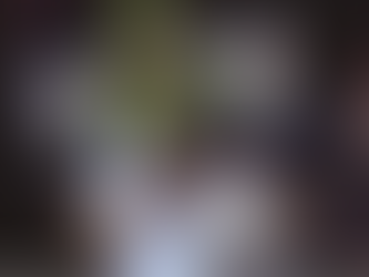 Two people in helmets hold flashlights in a dark room, with a green ghost-like figure smiling behind them. The mood is eerie yet playful.