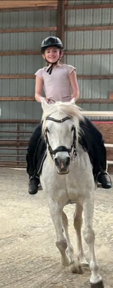 Girl in a helmet riding a white horse inside an arena, smiling at the camera.