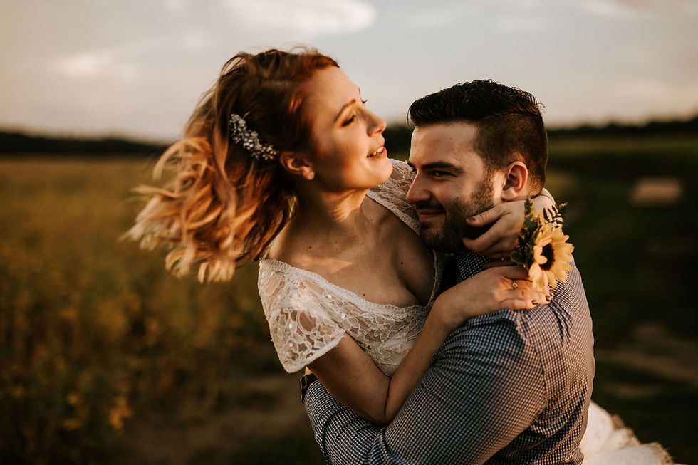 Couple embracing in a field at sunset. The woman wears a lace dress and sunflower corsage, smiling joyfully. Warm, romantic mood.