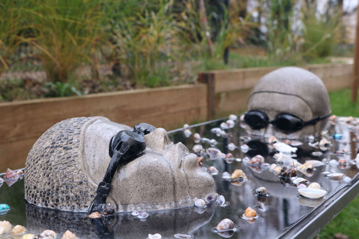 Installation artistique représentant deux têtes sculptées en céramique avec un bonnet de bain et des lunettes de natation, partiellement immergée dans l'eau avec des coquillages. Œuvre contemporaine interrogeant la montée des eaux et notre rapport à l'environnement. Art engagé, sculpture environnementale, changement climatique.