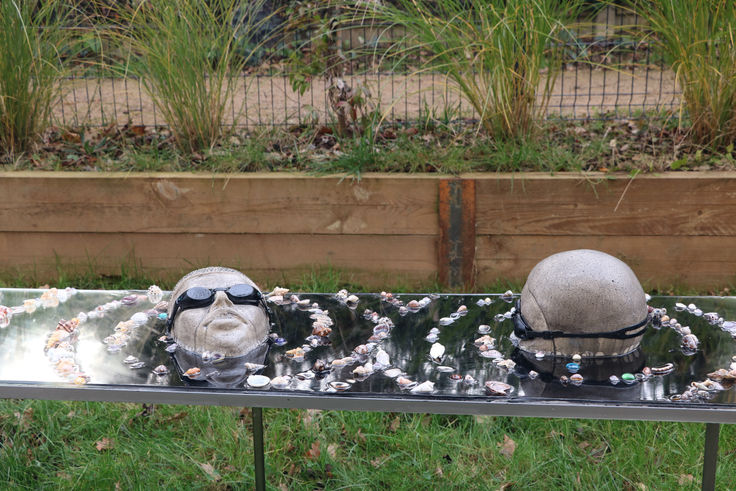 Installation artistique composée de têtes sculptées en argile, partiellement immergées dans une surface noire mémoire réfléchissante, entourées de coquillages. Cette œuvre questionne la montée des eaux, la des océans et notre rapport à l'environnement. Art engagé, sculpture contemporaine, changement climatique, installation immersive.