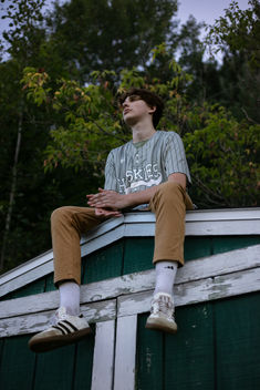A high school boy with dark wavy hair wearing his baseball jersey sitting on the roof of a weathered building for his senior photos at Franklin Academy Malone, NY