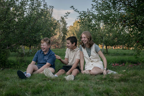 Three older siblings sit in an apple orchard in the fall and laugh for a family photo in Lake Placid NY