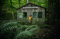 A high school girl wearing a yellow shirt and jean shorts stands against a sugar shack cabin behind some ferns for her senior photos in Malone NY