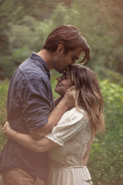 Engaged couple in a forest with golden light in Plattsburgh NY during early Summer. Him with wavy dark hair wearing a button up long sleeve blue shirt and her with long brown hair wearing a white cottage dress he kisses her forehead