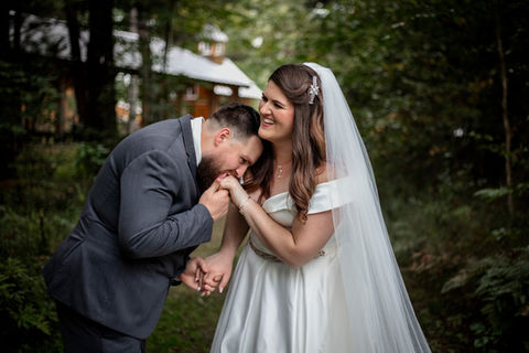 A tender moment of the groom kissing his bride's hand as she laughs, set in a forest in early fall.