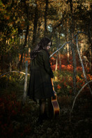 A girl walks away with her guitar for her senior photos in fire red foliage during sunset wearing a short black dress and long black jacket