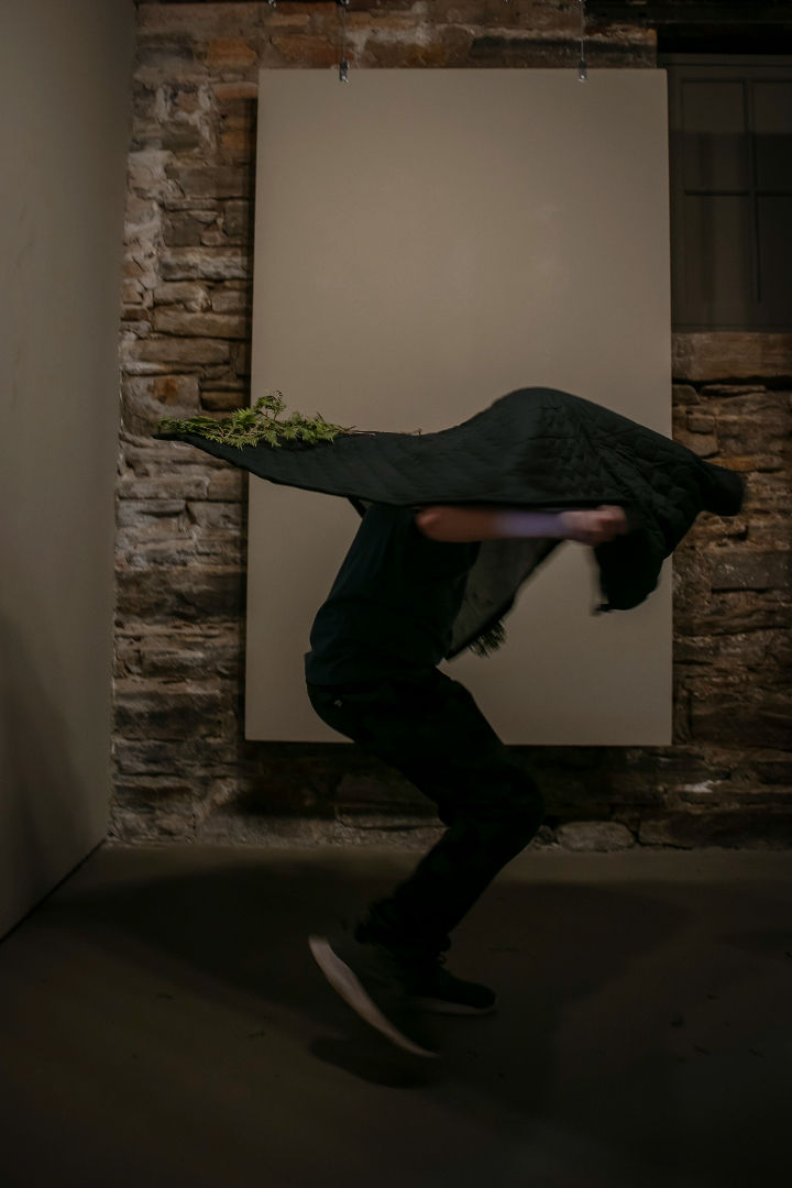 Person holding a dark quilted fabric with plants, bending forward in a dimly lit room with stone walls, creating a dynamic motion.