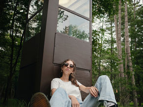 A high school girl with brown hair and sunglasses sitting against an old phone booth with sunglasses at Meacham Lake in Malone NY for her senior photos