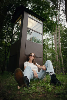 A high school girl with brown hair and sunglasses sitting against an old phone booth with sunglasses at Meacham Lake in Malone NY for her senior photos