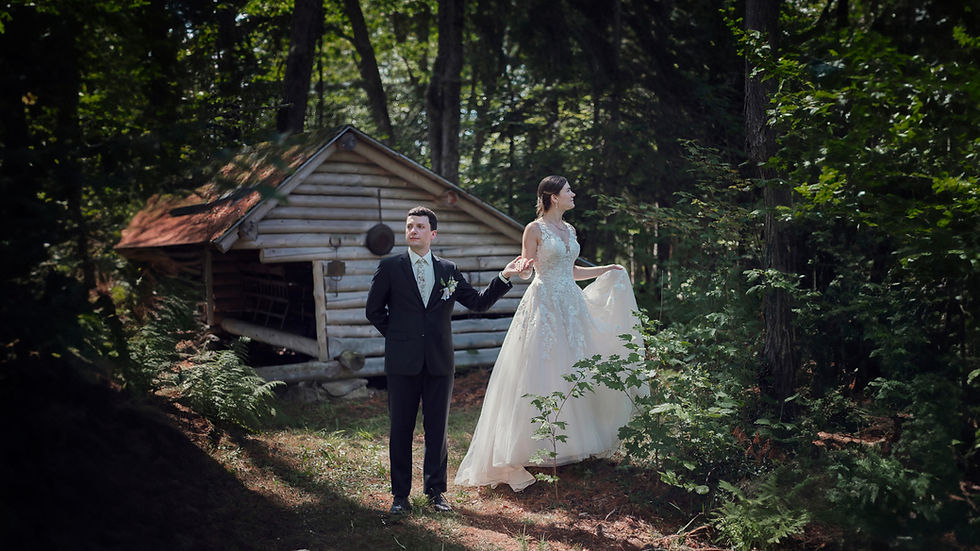 A bride stands on a tree stump holding her groom's hand in an Adirondack Coast forest with a leanto in summer_