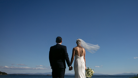 A romantic shot of a bride and groom holding hands, her veil flowing in the air, overlooking Lake Champlain in late summer.