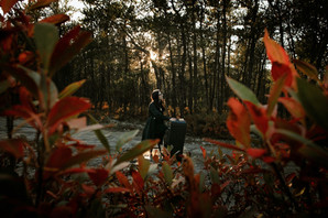 A high school girl plays her piano for her senior photos in fire red foliage during sunset wearing a short black dress and long black jacket.