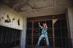 A high school boy with long blonde hair wearing a green Hawaiian shirt and jeans climbs an iron window in an abandoned building for his senior photos near Malone, NY