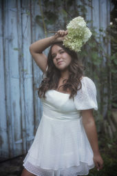 A high school girl with long dark hair wearing a short white dress holds a white bouquet of flowers over her head standing in front of a weather blue barn for her senior photos near Malone NY