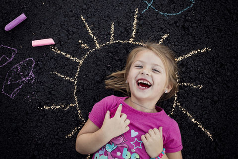 Little girl in pink shirt lays on the sun she made with chalk in her driveway while she laughs