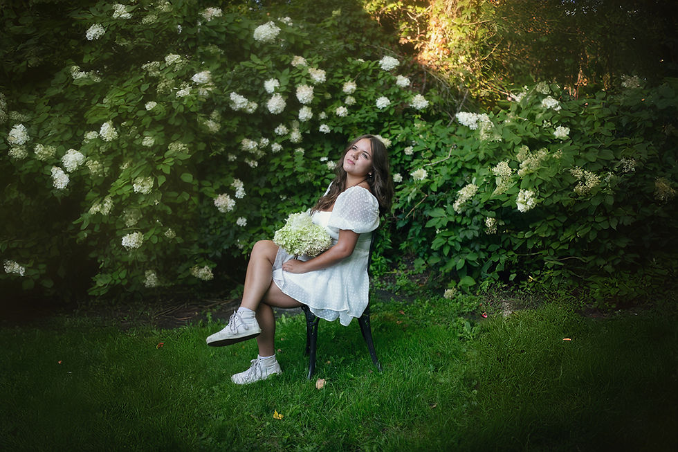 Woman in white dress sits on a chair in a garden, eyes closed, holding hydrangeas. Greenery and white flowers in the background. Relaxed mood.
