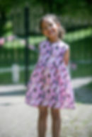 Little girl smiling in a park, wearing a pink dress with Barbie dolls printed on it.